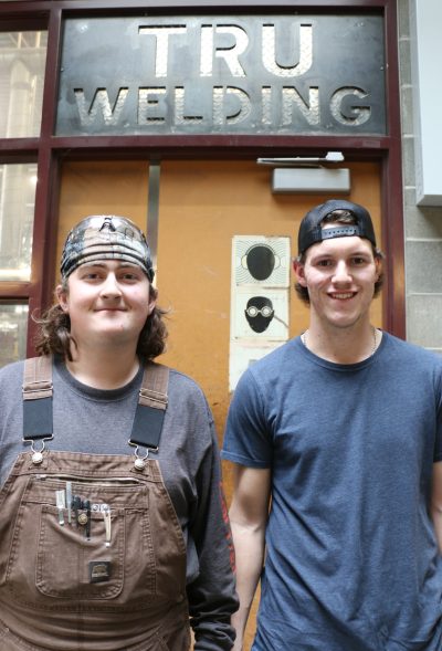 Two young men stand in front of TRU Welding sign.