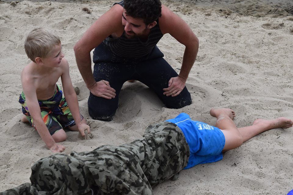 An instructor kneels beside children on the beach showing them how help an unconscious person rescued from the water.
