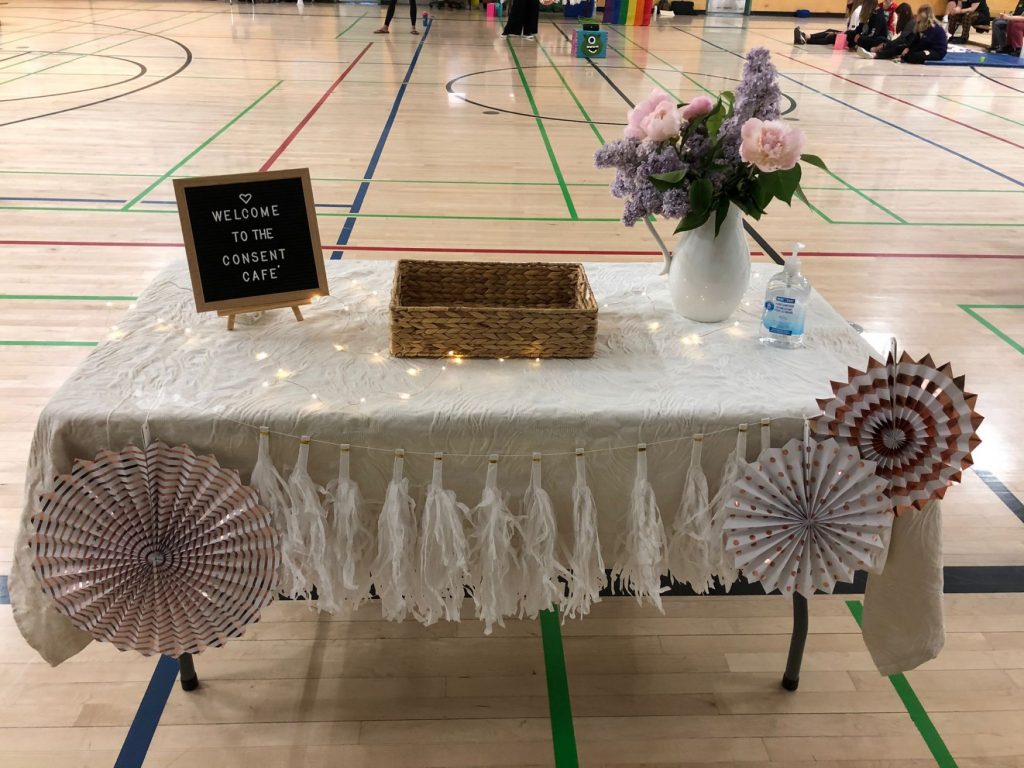 Table covered in a white cloth, decorations, a "Welcome to the Consent Café" sign, and a vase with pink flowers. 