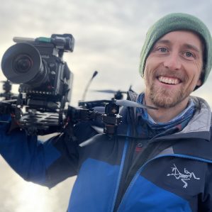 Man smiling at the camera while holding a large video camera.