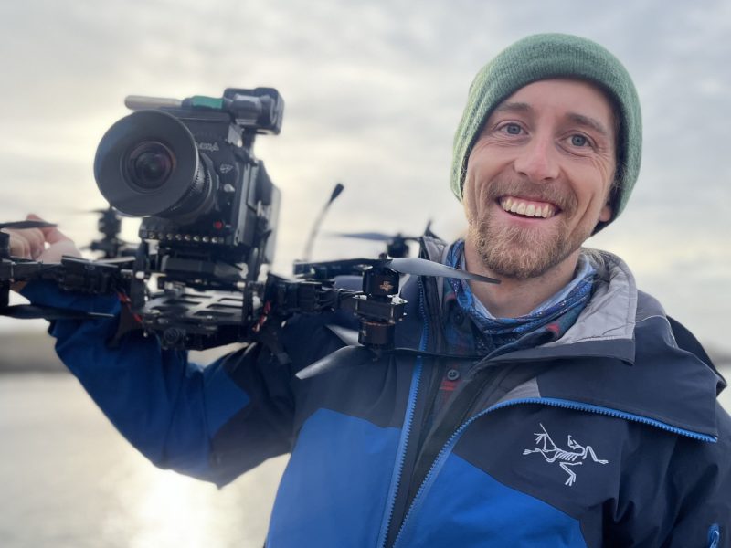 Man smiling at the camera while holding a large video camera.