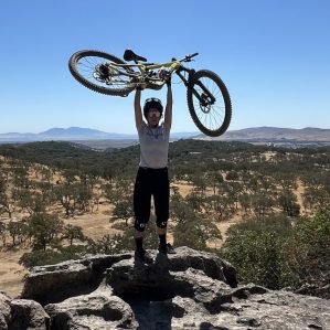 Woman holding a mountain bike in the air a top a large rock.